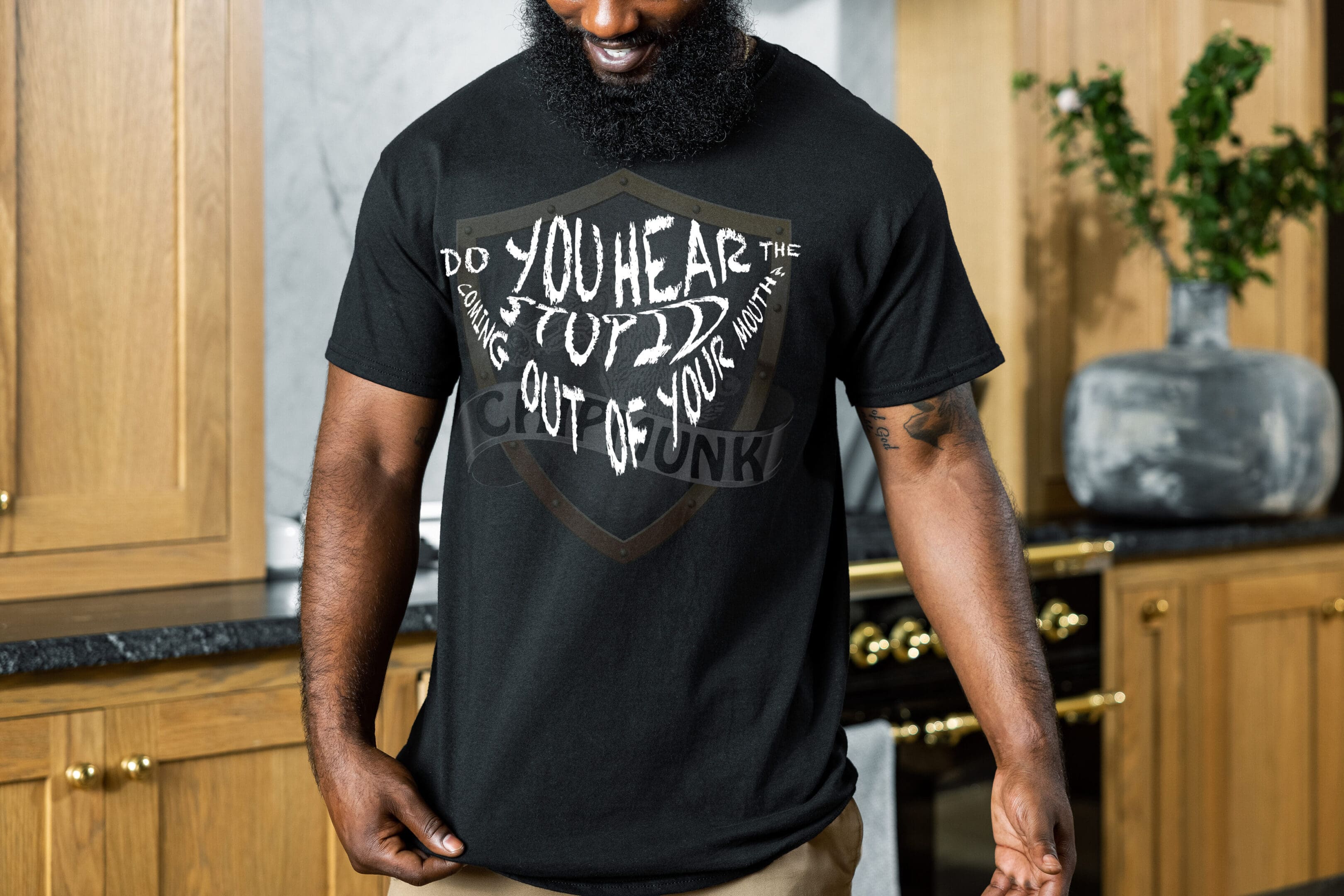 A man in a kitchen is wearing a Hear The Stupid T-Shirt, which is black with the text "Do you hear the stuff coming out of your mouth?" written on it in a decorative style.