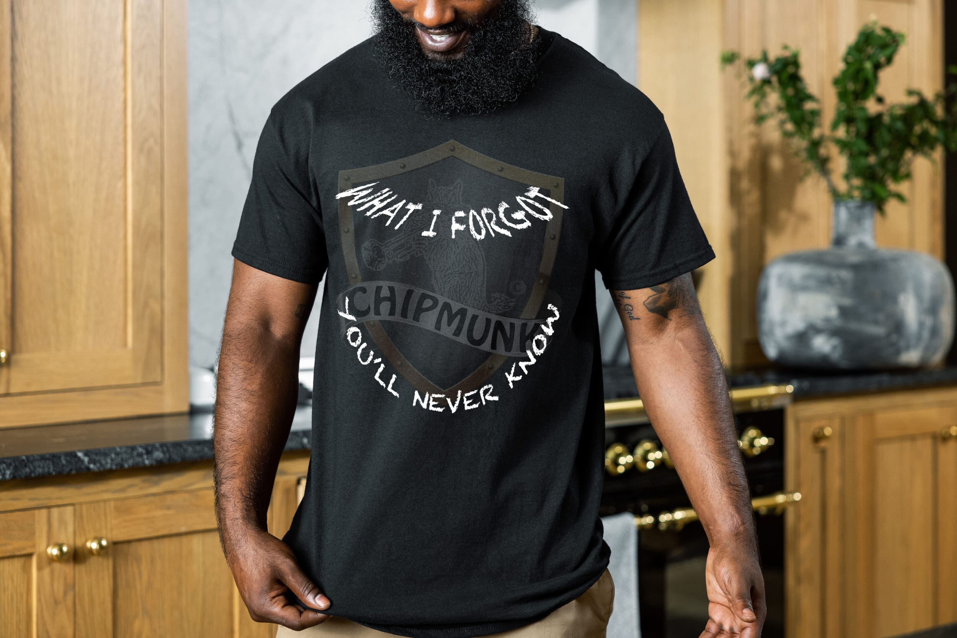 A bearded man in a kitchen wearing the "What I Forgot You Will Never Know T-Shirt," which is black with graphics including a shield and the text, "What I forgot, Ichipmunk, you’ll never know."