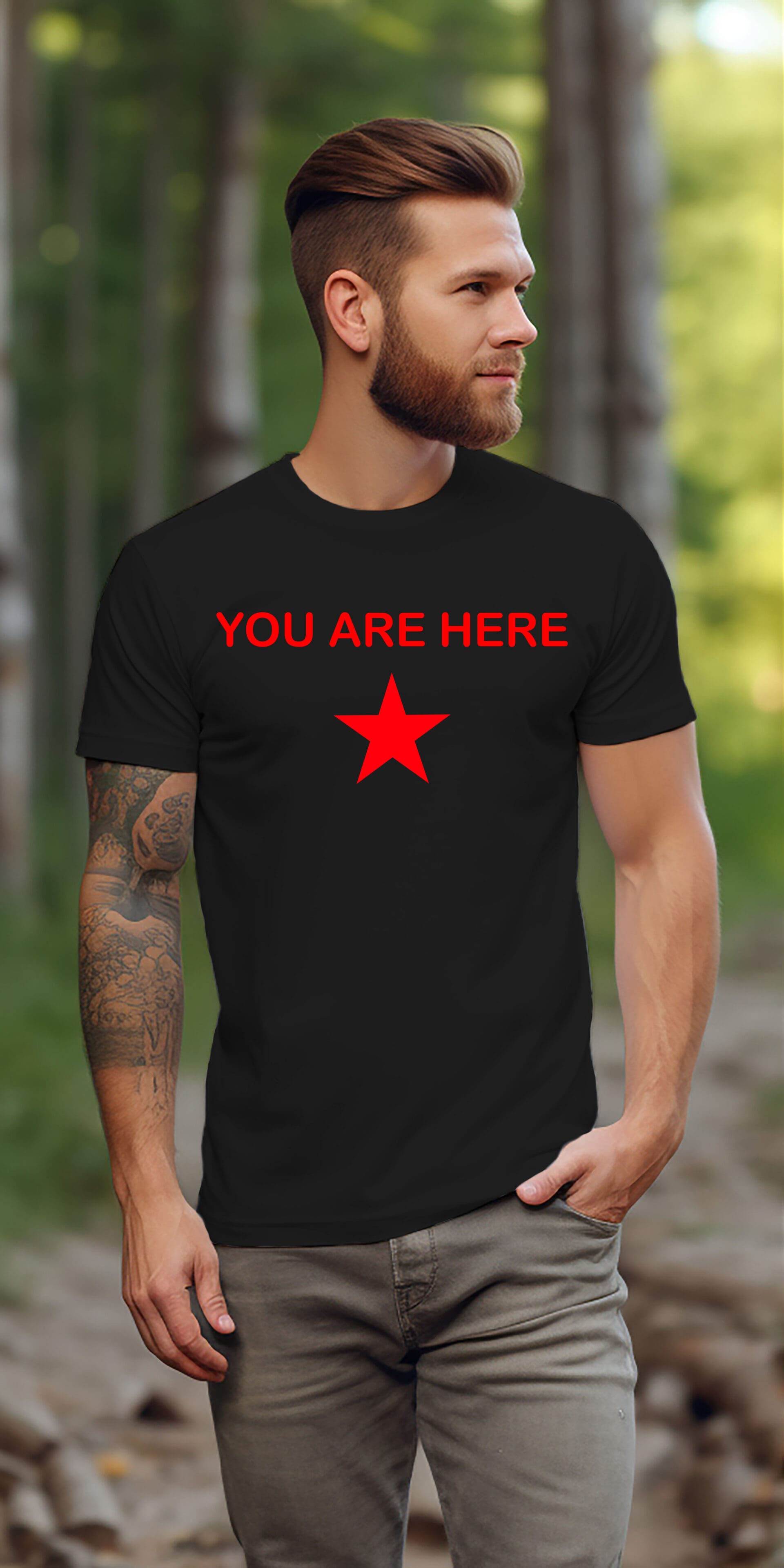 A man with a beard and tattoos stands outdoors wearing the Guiding Star Tee Shirt, which is black and features the text "YOU ARE HERE" above a red star.