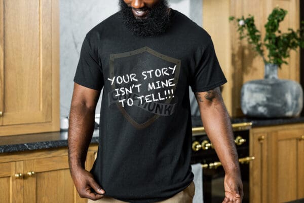 A person with a beard is wearing a black "Your Story Is Not Mine to Tell" T-shirt and standing in a kitchen.