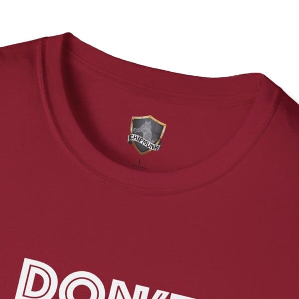 Close-up of a red t-shirt with "DONKE" partially visible in white letters. The inside label features a chipmunk logo embedded with text from the Don't Be A... Tee.