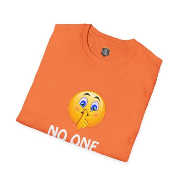 Folded orange t-shirt featuring a smiley face emoji holding a finger to its lips, accompanied by the text "No One Cares.