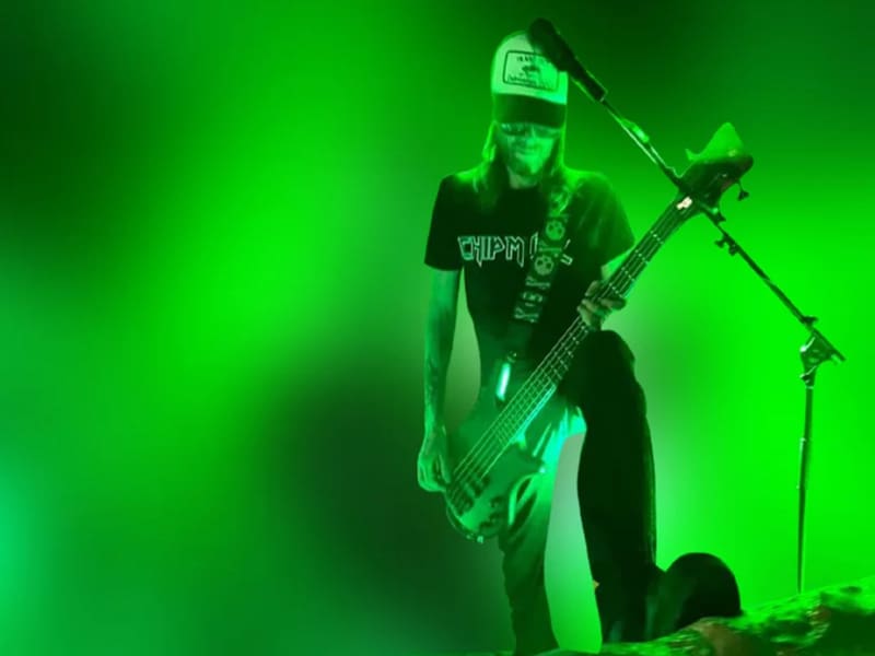 A musician wearing a cap plays a bass guitar on a stage with bright green lighting.